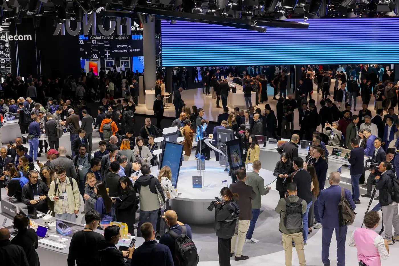 A large exhibition hall bustling with attendees at a major technology event. Various booths showcase innovative products, with digital displays and interactive setups drawing crowds. The scene includes tech enthusiasts, business professionals, and media personnel exploring the latest advancements in mobile technology, AI, and telecommunications. Bright signage and branding from companies like 'HONOR' and 'SK Telecom' are visible, reinforcing the high-tech atmosphere.